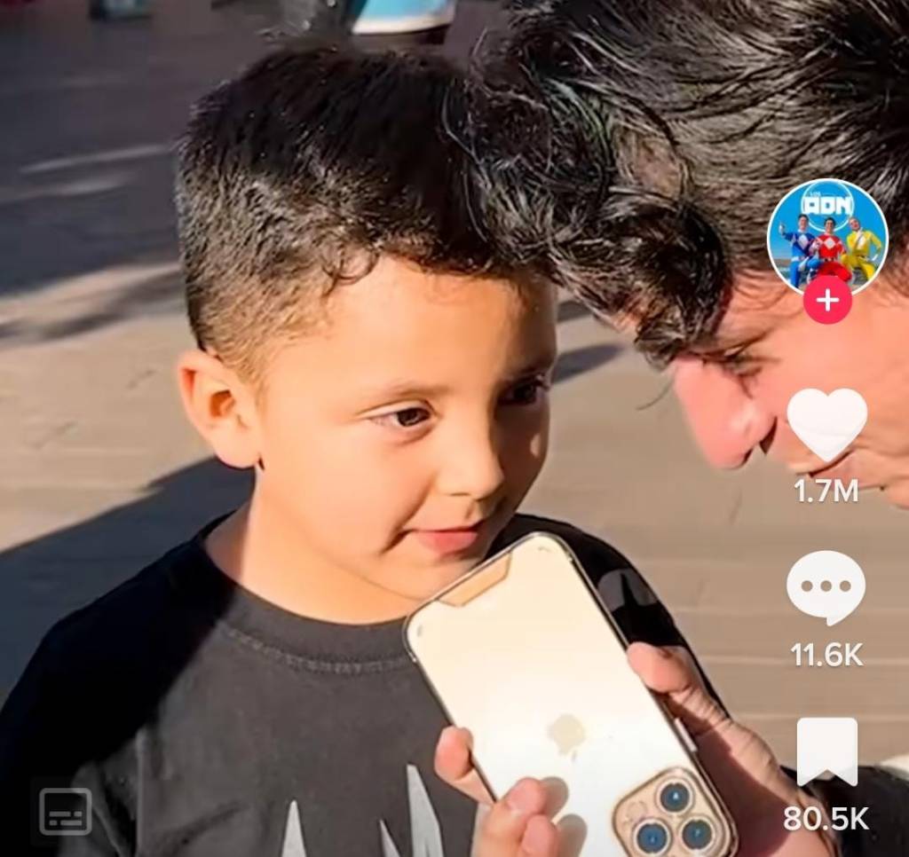 menino conta que pai trai mae em entrevista menino, por volta de 6 anos, falando com um entrevistador que segura um celular. Ambos são brancos e de cabelo castanho. Estão na rua, no sol.