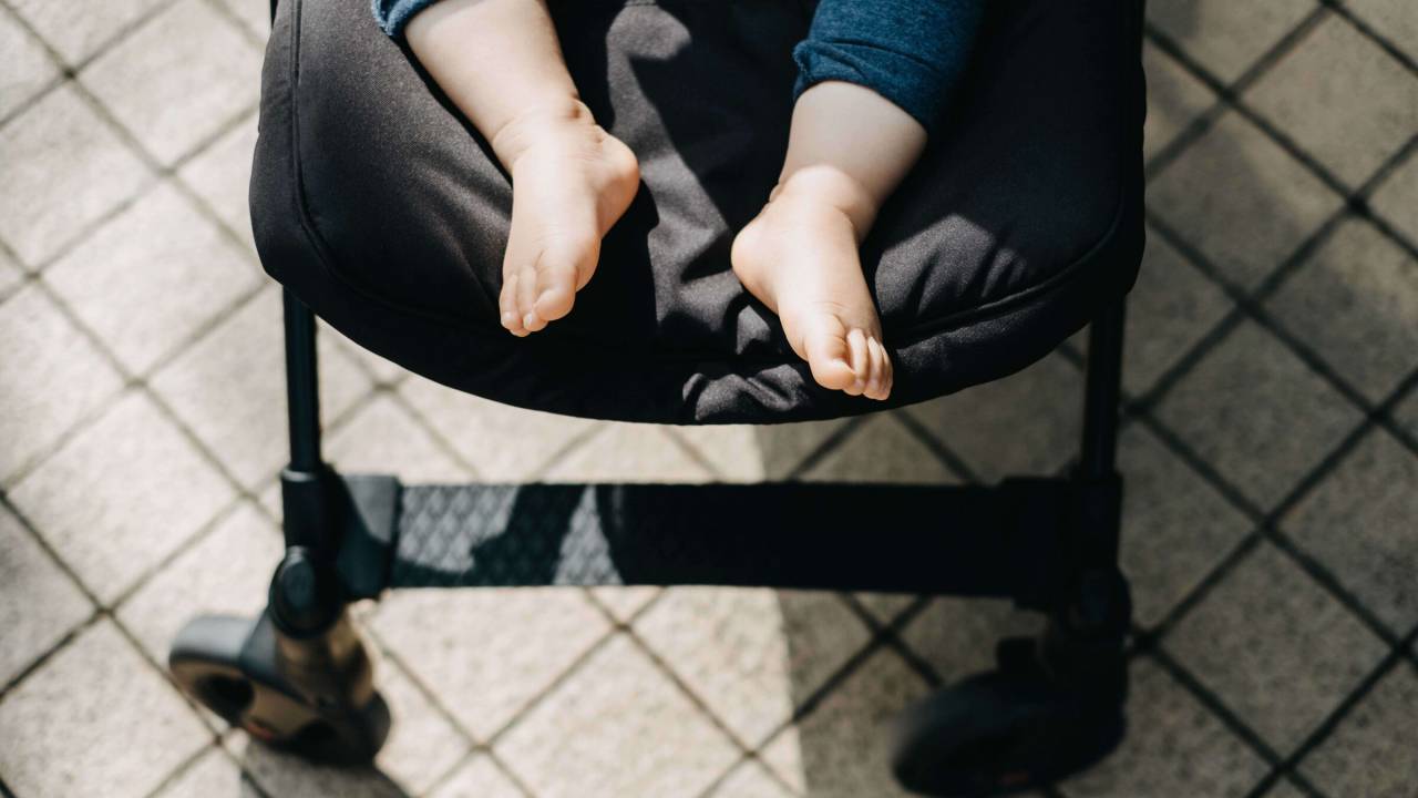 Parte mais inferior de um carrinho de bebê preto, aparecem apenas os pezinhos descalços da criança de pele branca. O chão é cinza.
