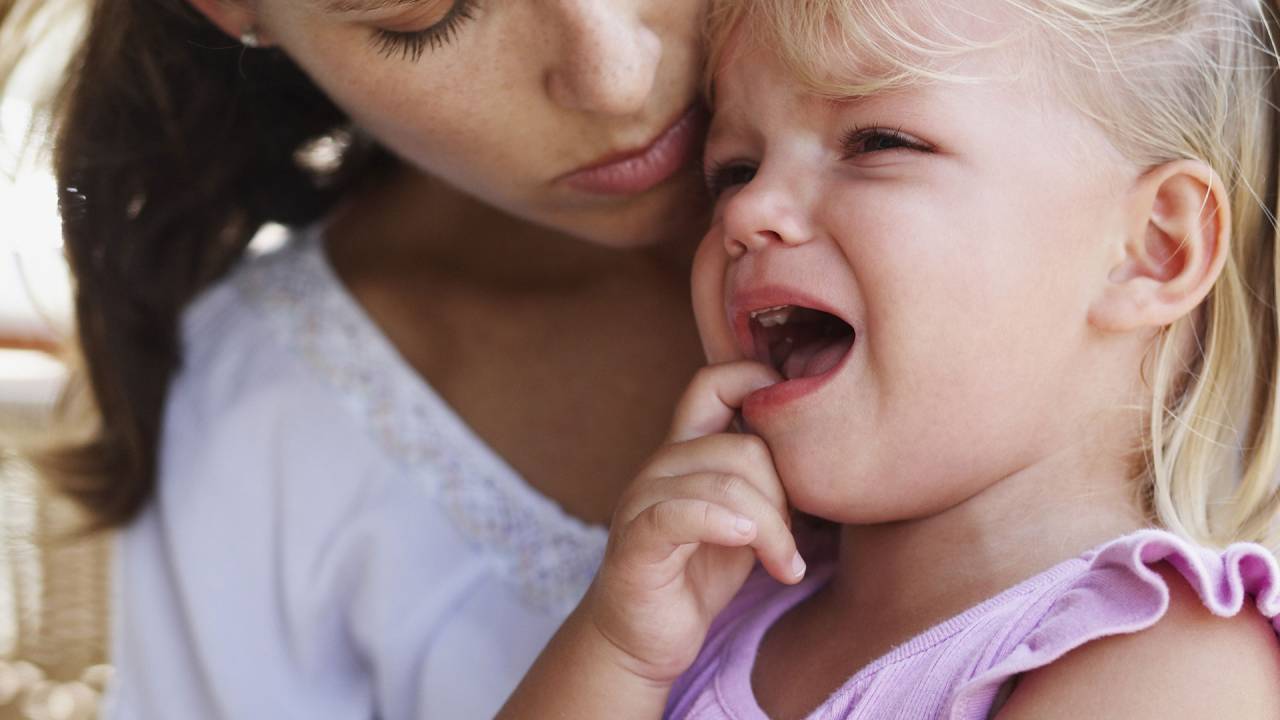 garota sentada no colo de uma mulher. A criança chora, colocando um dos dedos na boca