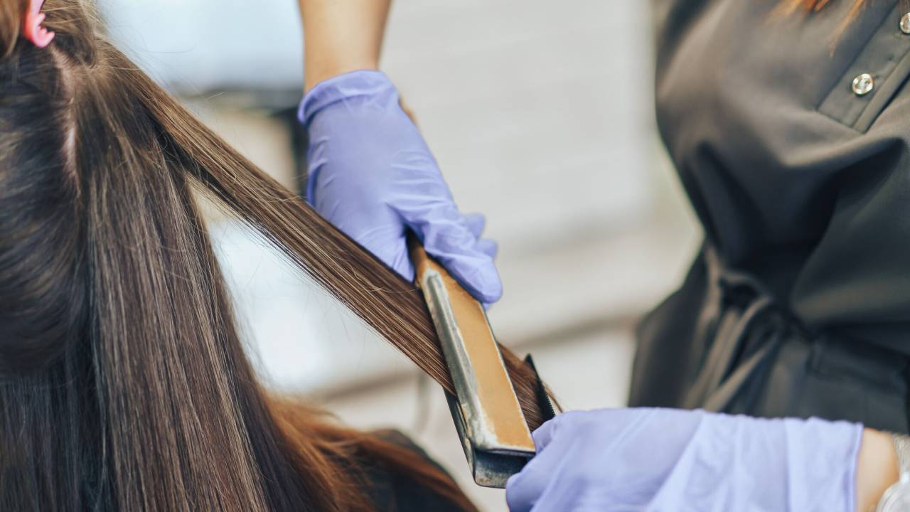 Mulher de cabelo comprido tendo o cabelo alisado por chapinha no salão de beleza. A cabeleireira usa luvas azuis.