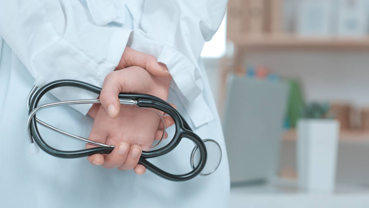 Médica com as duas mãos para trás, segurando um estetoscópio. Ela está de jaleco branco.