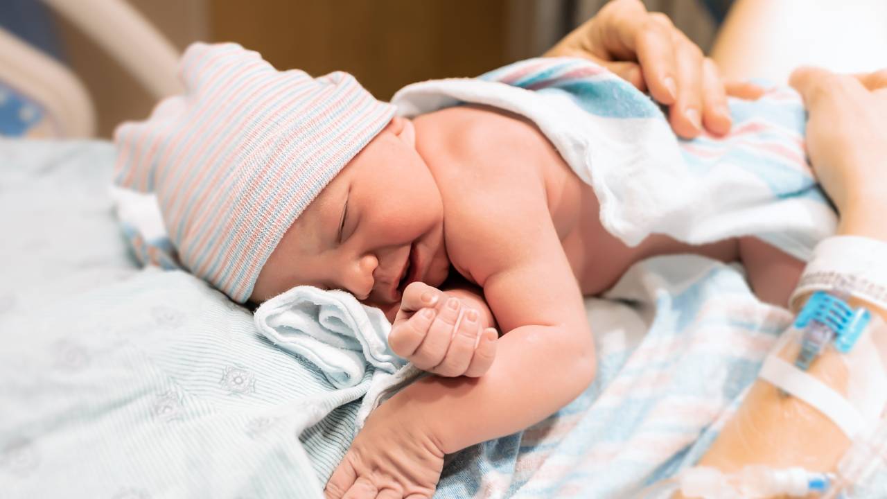 Bebê recém-nascido sobre o corpo da mãe no hospital. Ele usa uma touquinha, está sem roupa, apenas coberto por um lençol.