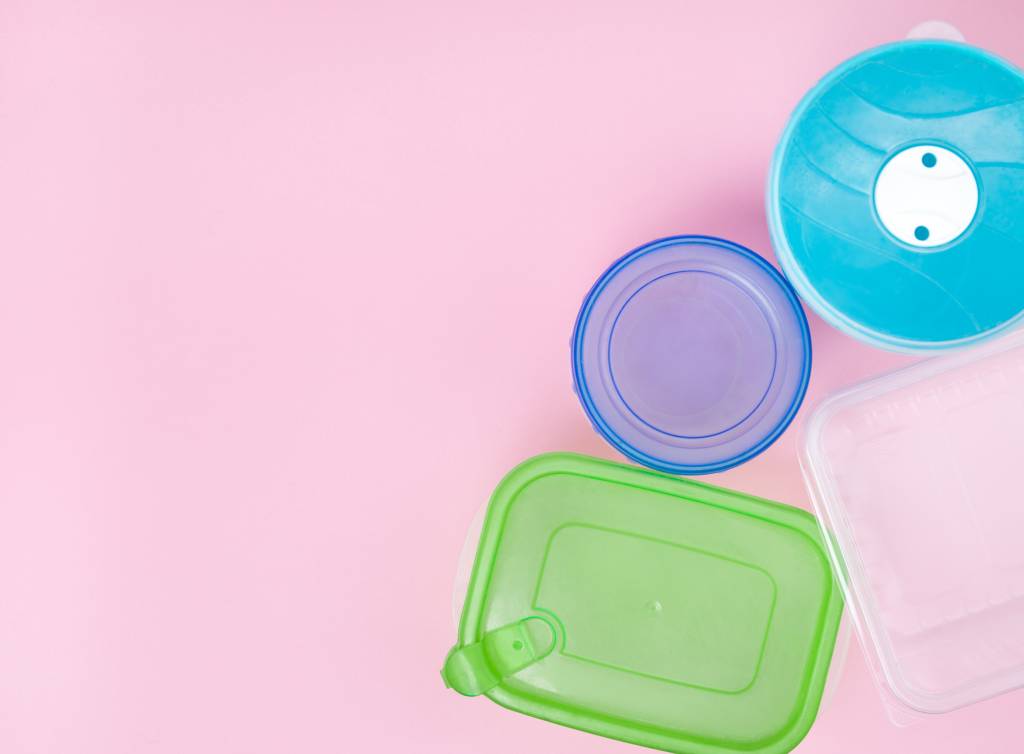 Potes de plástico para armazenar comida, com tampas coloridas (sobre fundo rosa claro).