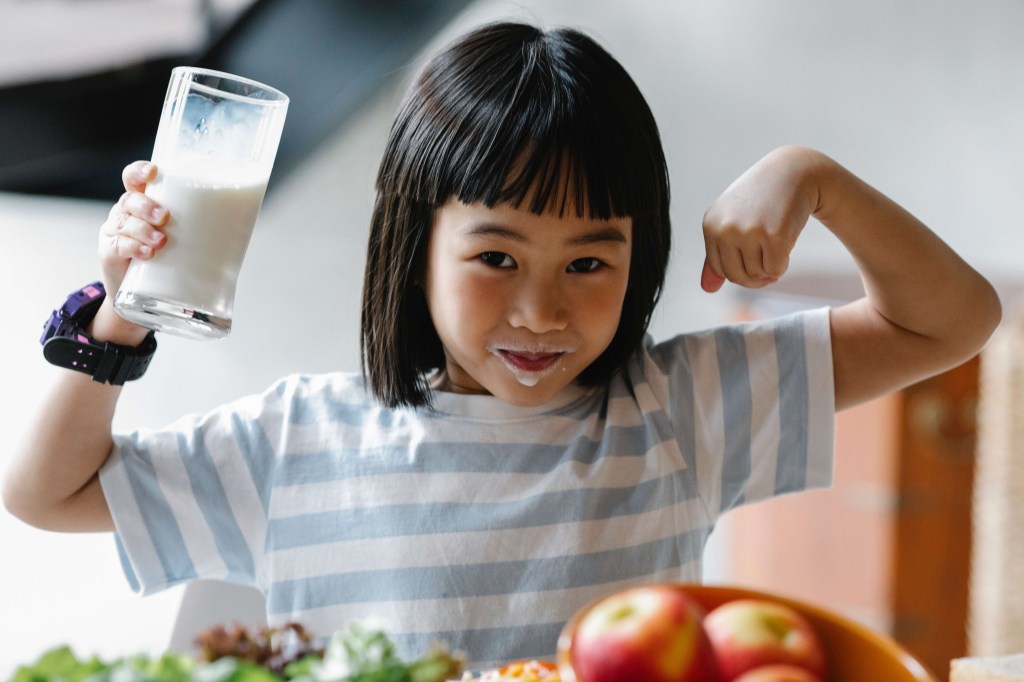 Menina asiática de cabelos pretos e franja com a boa suja de leite. Ela mostra a força com os braços para cima e segura um copo de leite com o outro. No primeiro plano, aparecem maçãs e verduras.