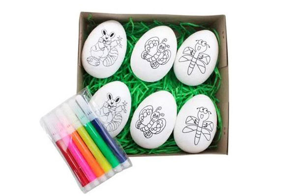 caixa de papel aberta. Dentro dela estão seis brinquedos em formato de ovo com desenhos para as crianças pintarem. Ao lado da caixa, há um conjunto de canetinhas coloridas