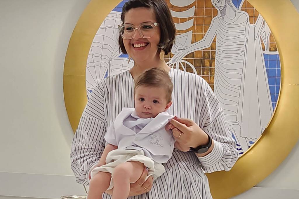 Tainá, mulher de roupa branca, pele clara, cabelos castanhos e curtos, carregando francisco, de 4 meses, também de pele clara e roupa branca. Estão na igreja.