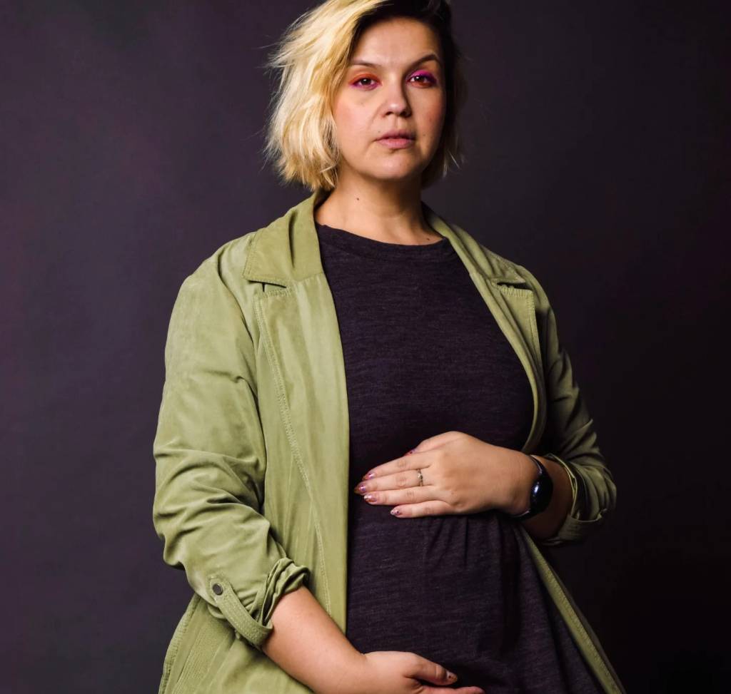 Tainá Goulart, mulher grávida, branca, usando vestido preto e camisa verde aberta por cima. Está com as duas mãos sobre a barriga. O fundo da foto é preto.