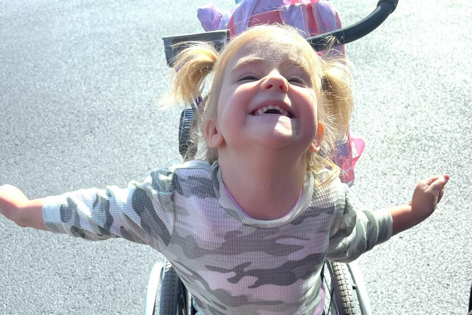 Blaire Spivey, criança pequena, de pele branca, loira, em cadeira de rodas, com os braços abertos e sorrindo para a foto.