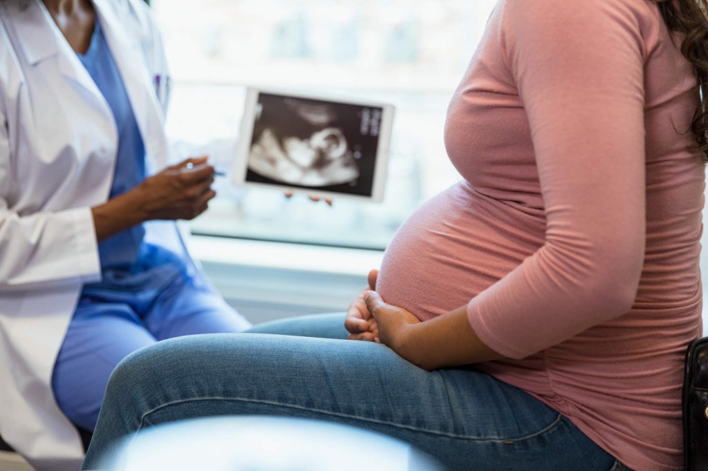 Mulher grávida sentada conversando com médica também sentada. A médica aponta para a imagem do bebê no ultrassom.