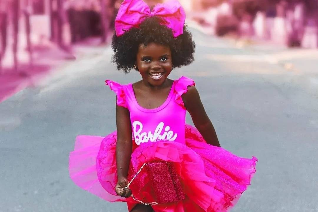 Criança vestida de Barbie, pulando e sorrindo no meio da rua. Ela é negra, tem por volta de 5 anos, usa collant, saia de tule, sandálias e laço na cabeça - tudo rosa pink.