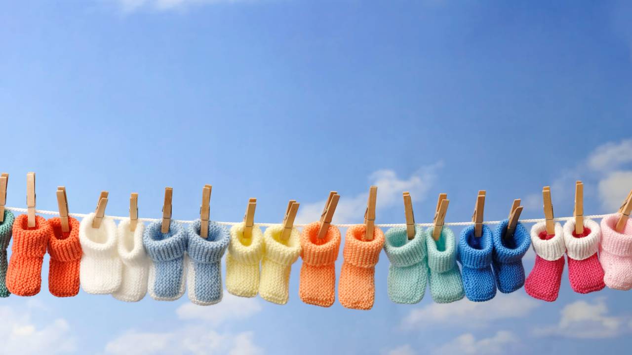 Diversos sapatinhos de tricô de diferentes cores pendurados em um varal. O plano de fundo é um céu azul com poucas nuvens brancas.