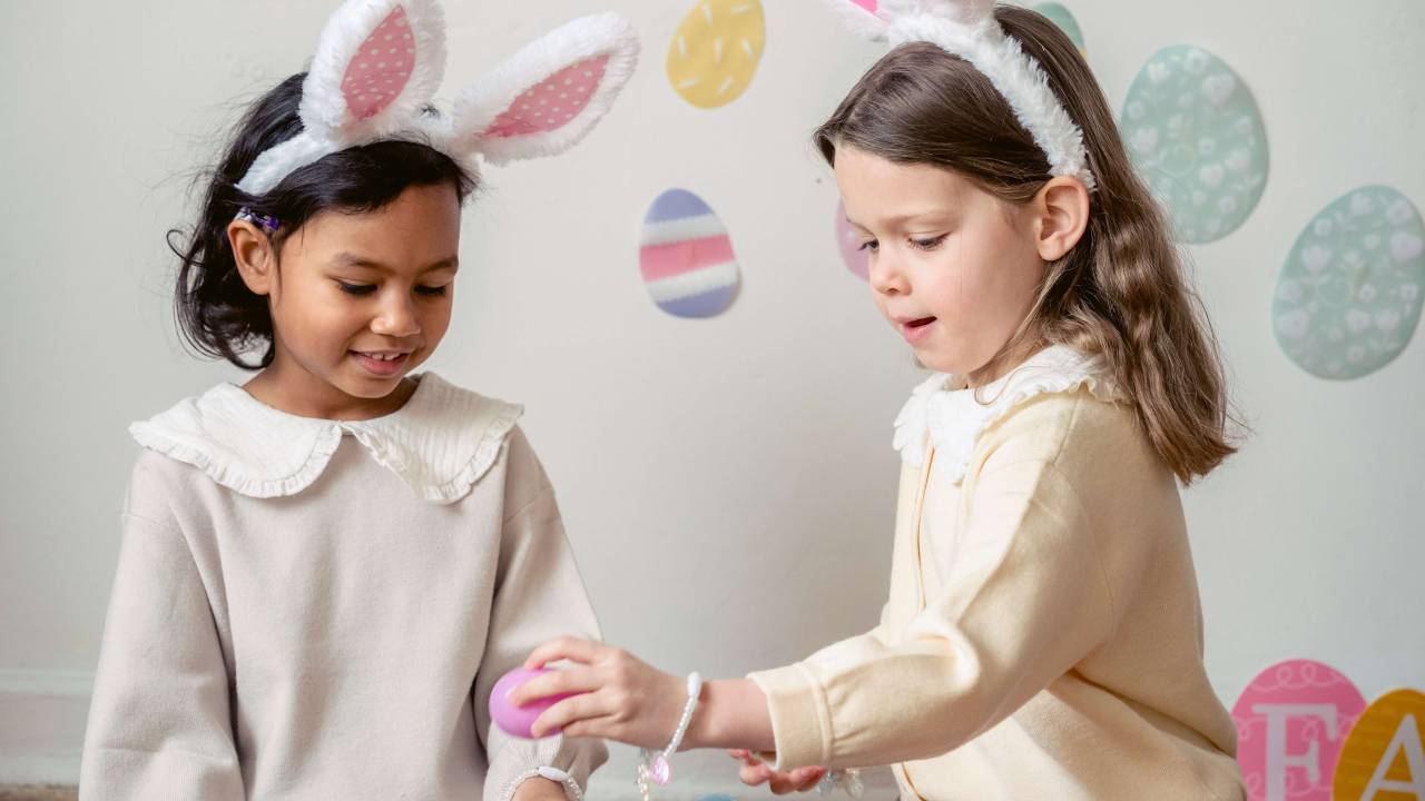 Duas meninas sentadas brincando com ovos coloridos. Elas usam orelhas de coelho.