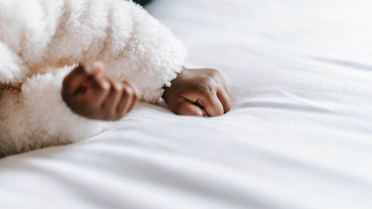 Bracinhos e mãozinhas de bebê negro deitado sobre lençol branco. É possível ver as mangas peludinhas brancas de uma roupa de inverno.