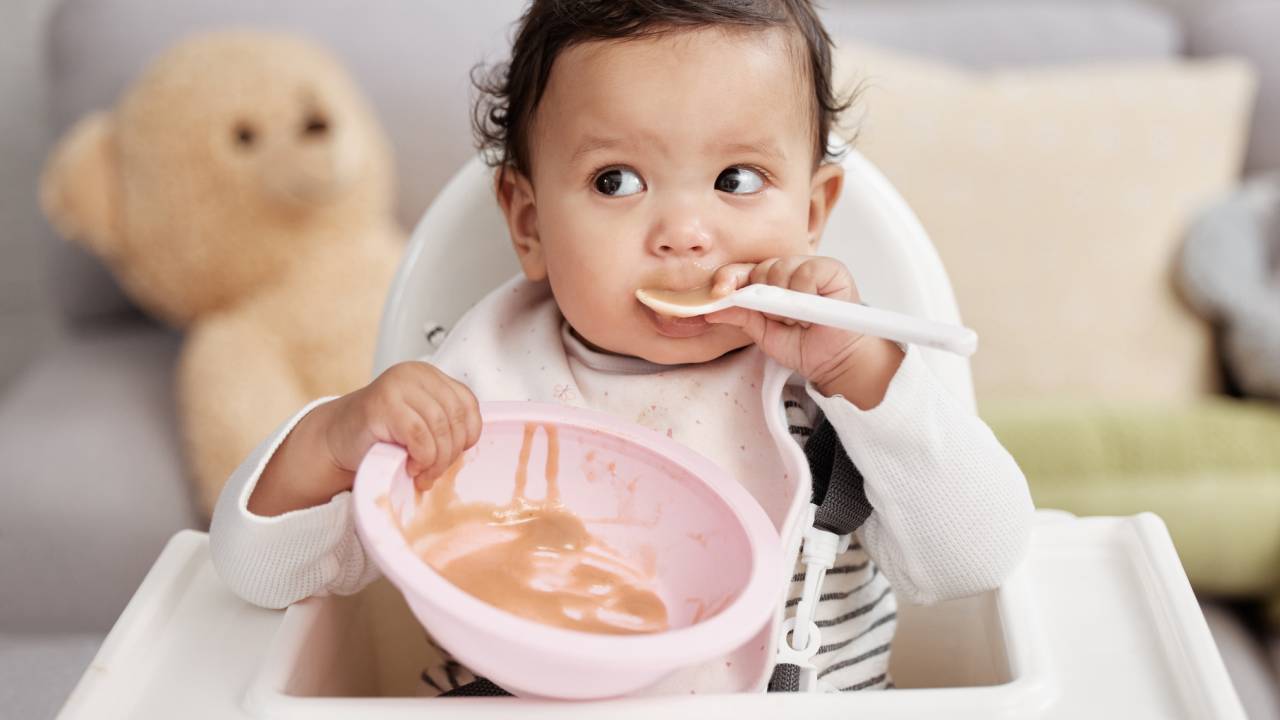Bebê comendo sozinho com a colher na boca e segurando o pote com a outra mão.