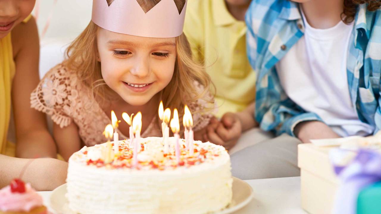 menina usando uma coroa de papel olhando para um bolo de anicersário cheio de velas e sorrido. Há outras pessoas ao redor dela