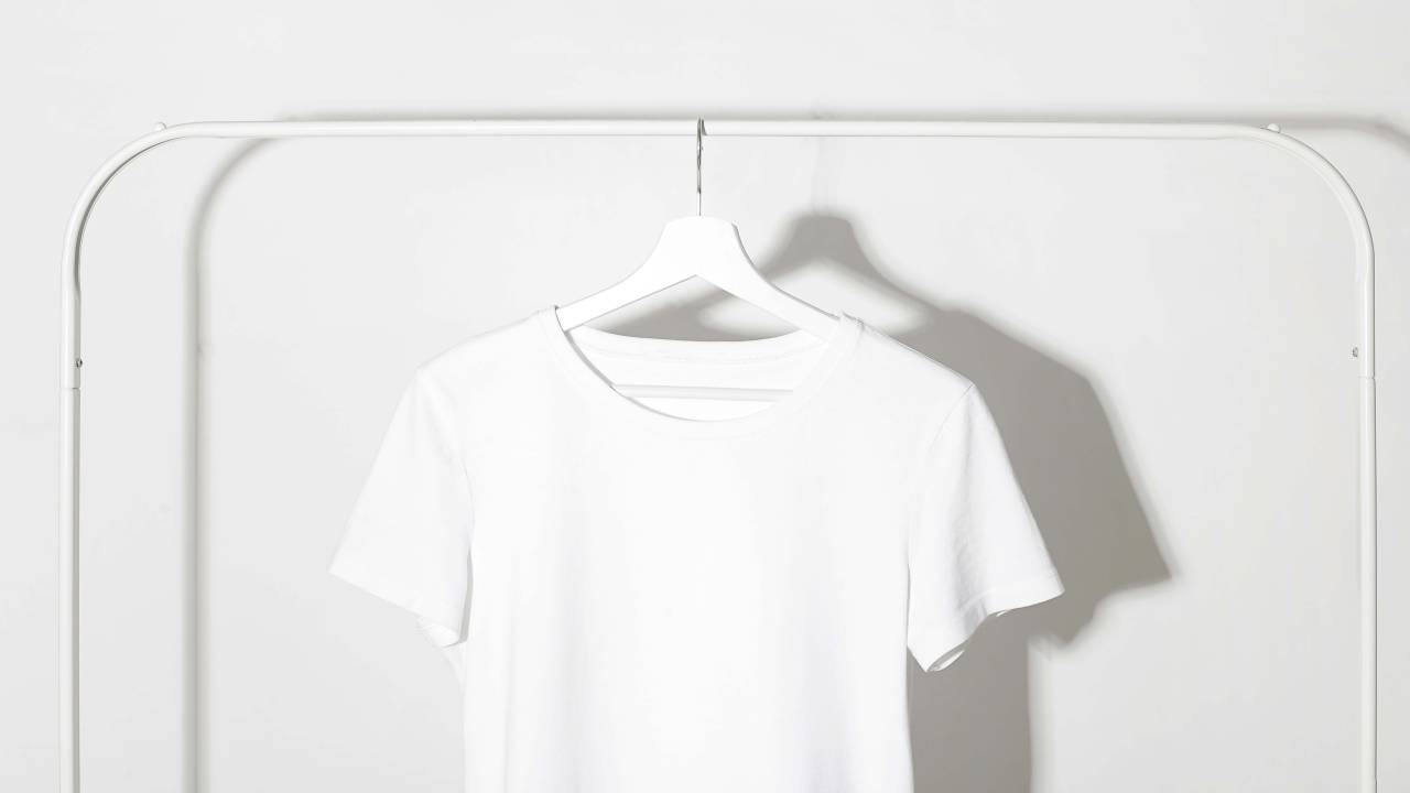 Camiseta branca em cabide branco pendurado em arara branca. A parede de fundo também é branca.