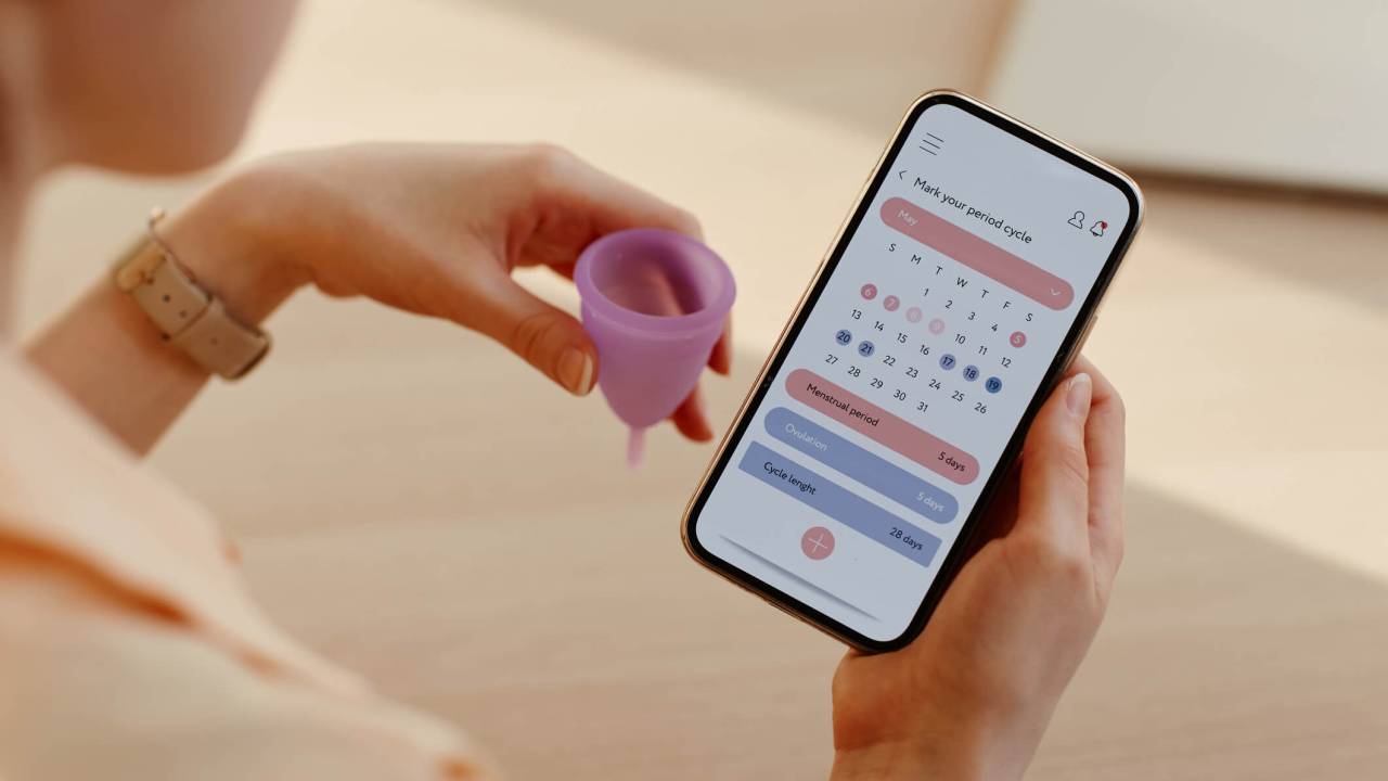 Mulher segurando o celular com a mão direita, onde olha um calendário menstrual, e um coletor menstrual rosa com a mão esquerda.