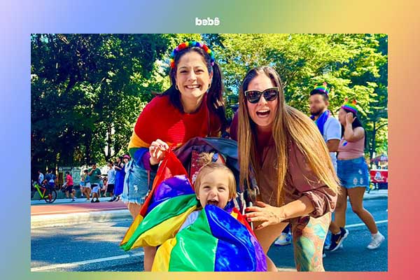 Duas mulheres inclinadas junto de uma criança, que segura a bandeira LGBTQIA+. Todos sorriem
