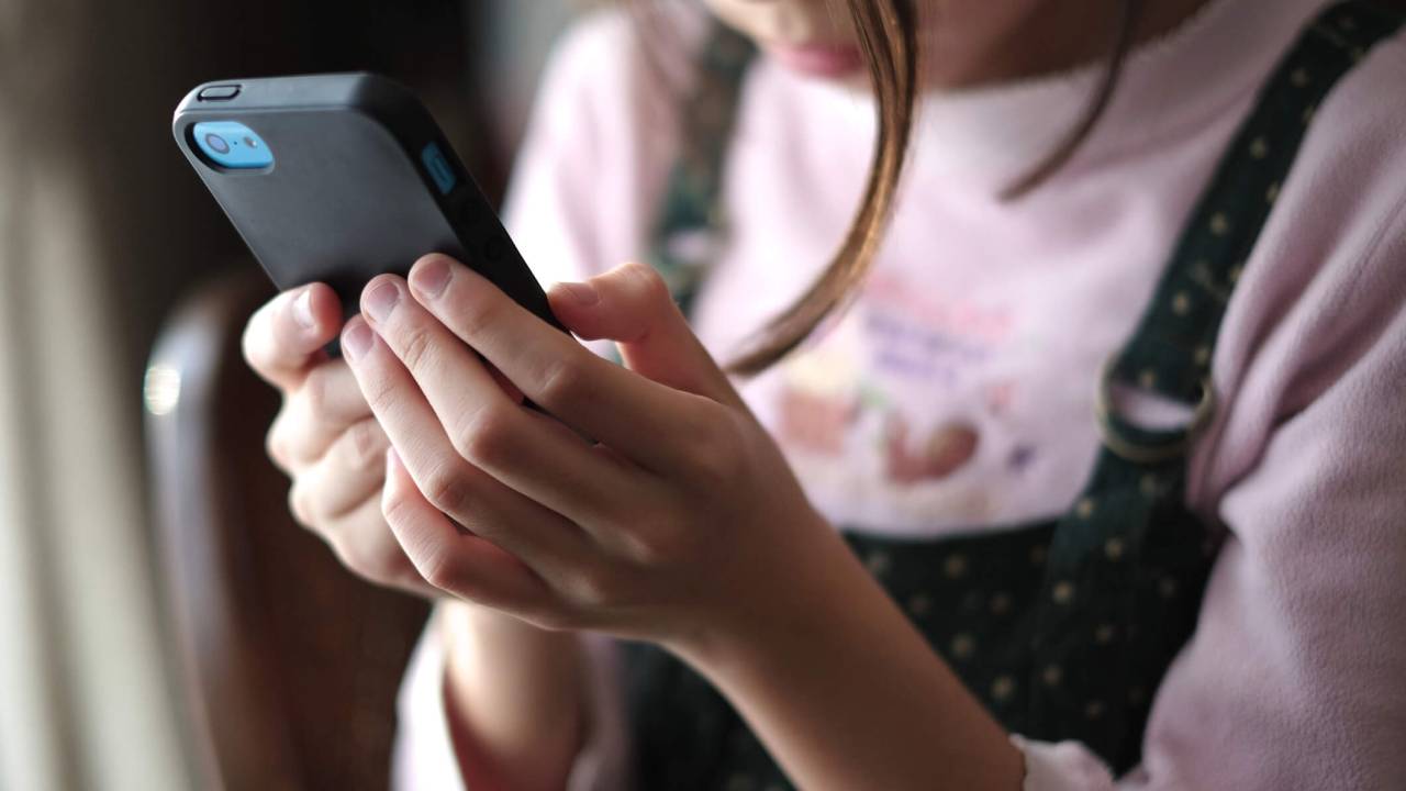Menina segurando smartphone com as duas mãos. Não é possível ver seu rosto, apenas parte dele e mechas de cabelo soltas.