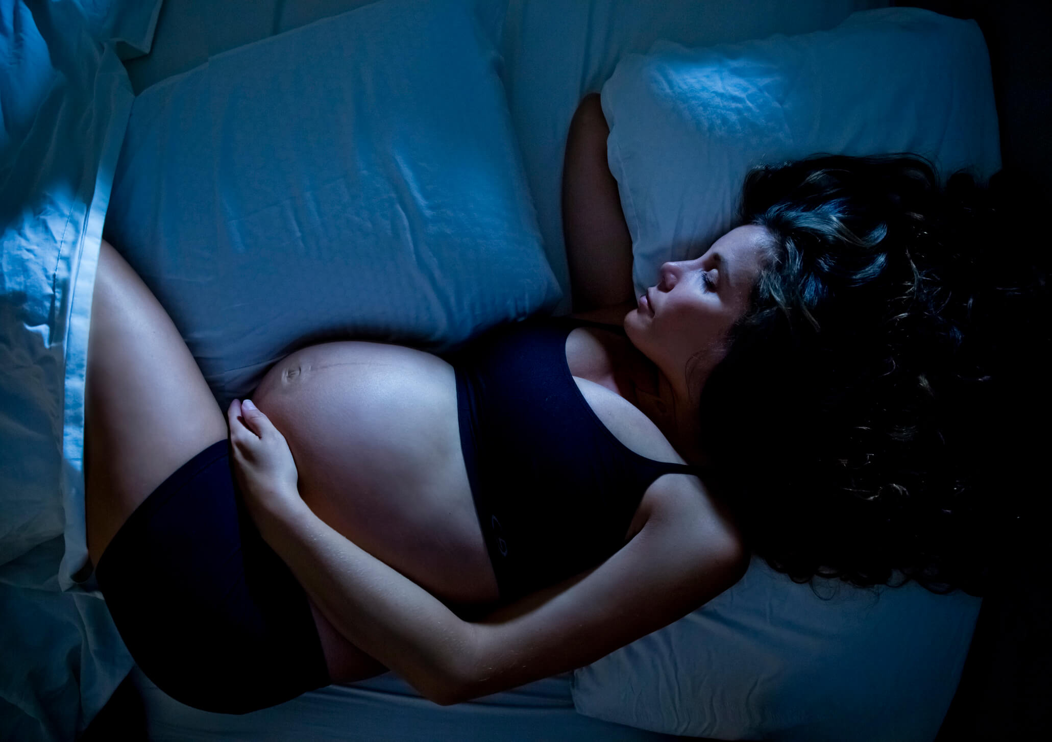 Mulher grávida dormindo na cama no escuro. Ela está deitada com um braço embaixo do travesseiro e outra na barriga. Está de top e shorts pretos.