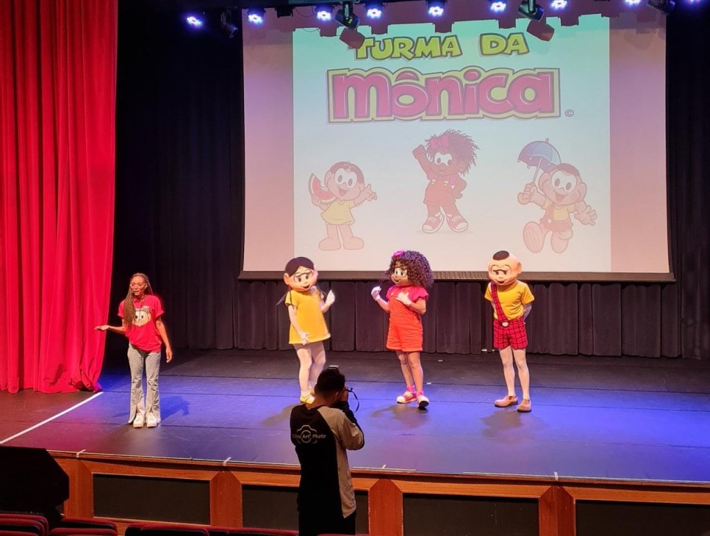 Personagens da turma da mônica no palco: Magali, Milena e Cascão. Há, também, uma moça monitora infantil