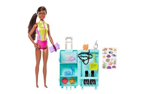 Barbie bióloga marinha. Ela usa roupa de mergulho e está ao lado de uma mala de viagem aberta, onde estão objetos como óculos de mergulho e pé de pato, além de peixe, estrela do mar e concha