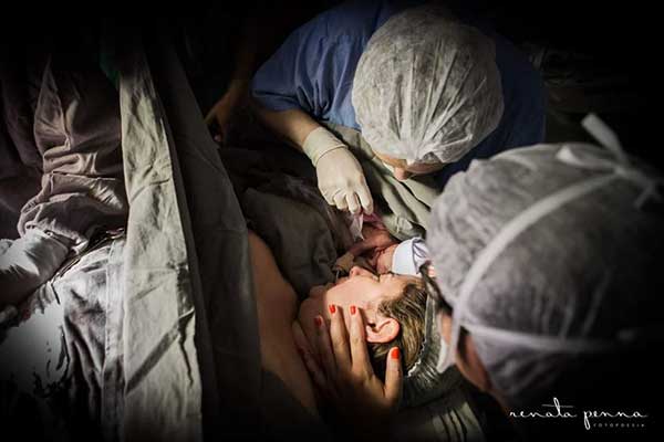 Mulher deitada em uma mesa cirúrgica com um homem e um bebê recém-nascido ao lado dela