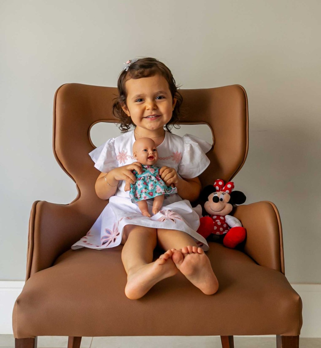 garotinha sentada em uma cadeira, segurando uma boneca e sorrindo para a câmera