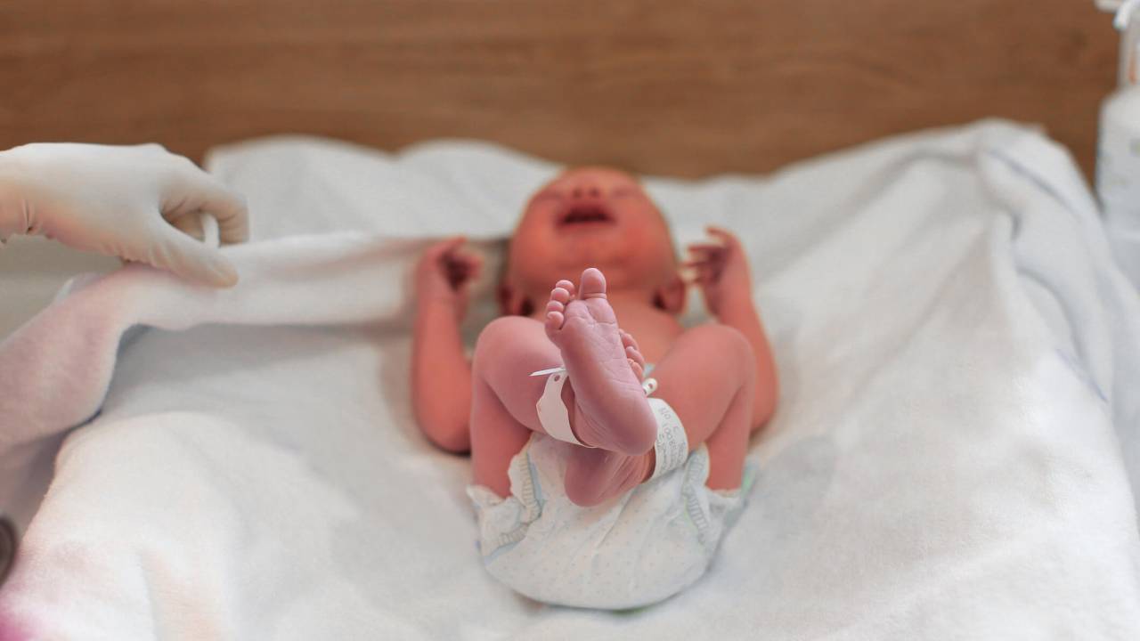 Bebê recém-nascido usando fralda, deitado sobre lençol branco, com uma pulseirinha de identificação em cada tornozelo.