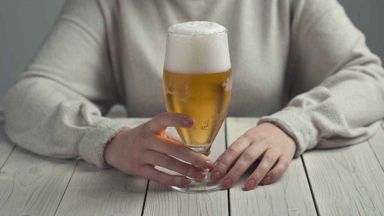 mulher sentada diante de uma mesa, com foco na mão dela segurando um copo de cerveja que está sobre o móvel