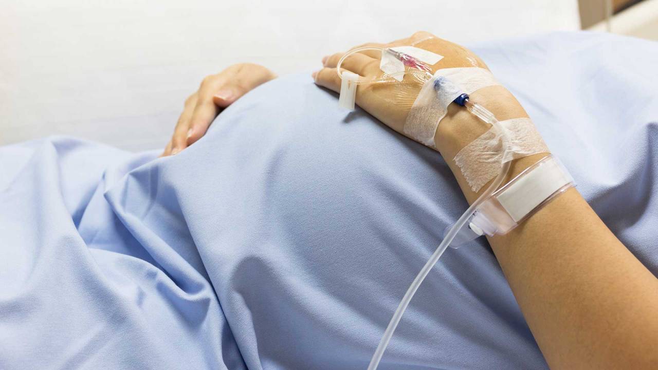 Foco na barriga de uma mulher grávida usando roupas de hospital. Ela tem um acesso em uma das mãos