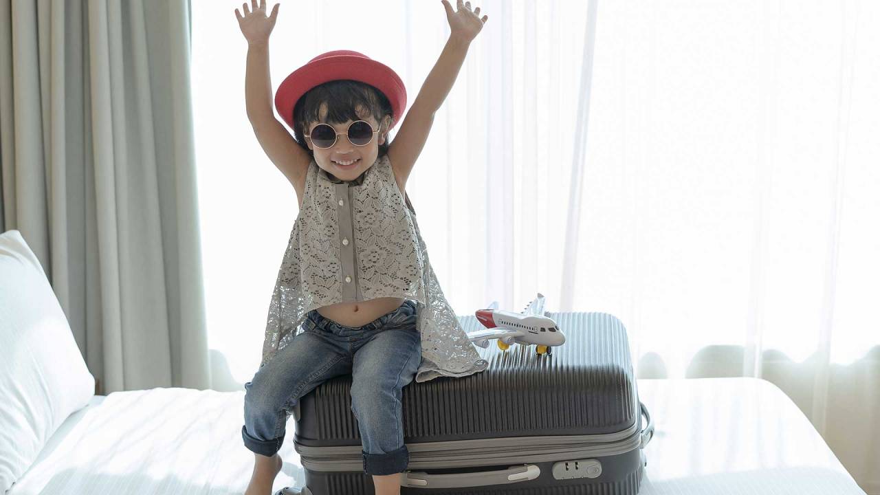 garotinha sentada sobre uma mala de viagem que está em cima de uma cama, em um quarto. Ela usa óculos escuros e chapéu, levanta os braços para cima e sorri