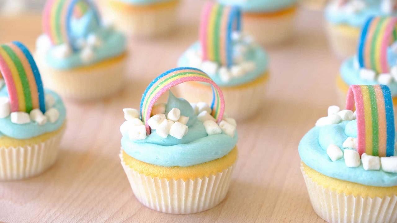 cupcakes enfileirados. Eles têm a fominha branca, cobertura azul com pedaços de marshmallows brancos e um enfeite em formato de arco-íris