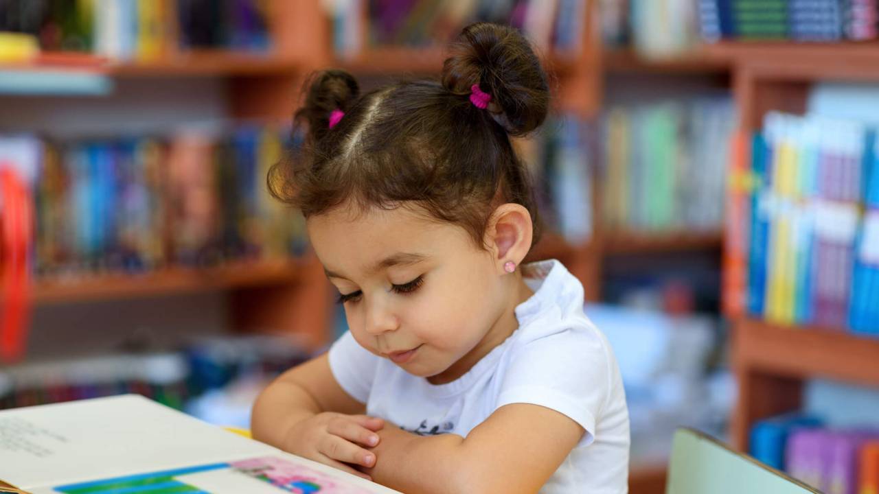 Menina de 2 ou 3 anos, sentada em livraria, lendo um livrinho apoiado em uma mesa amarela. Tem cabelos castanhos e presos em dois rabinhos no alto da cabeça.