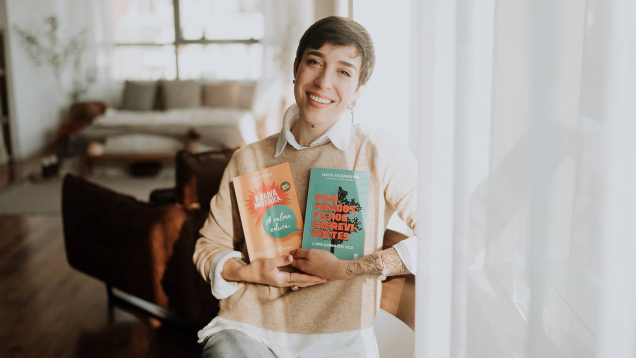 Maya é uma mulher branca, de cabelo castanho e bem curto. Está sentada, de jeans e blusa creme, segurando os dois livros em frente ao peito. Está sorrindo.