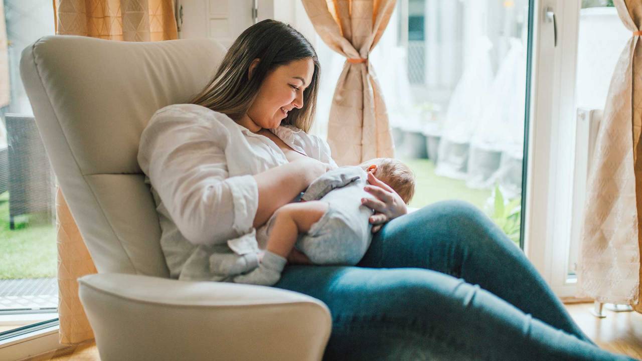 mulher sentada em uma poltrona de amamentação, diante de uma cortina, amamentando um bebê. Ela sorri para a criança