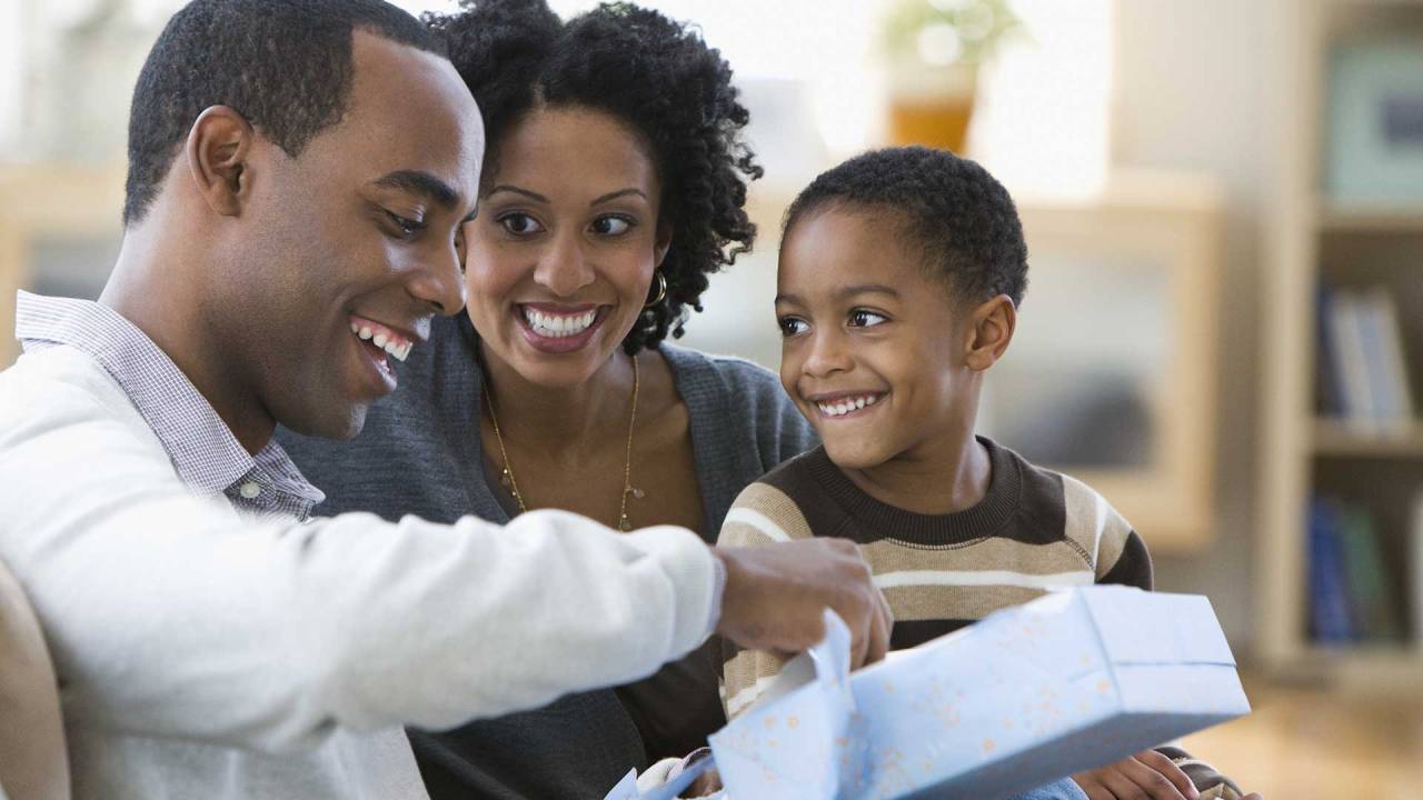 homem sentado, sorrindo e abrindo um presente enquanto uma mulher e um garoto, também sentados, olham para ele sorrindo
