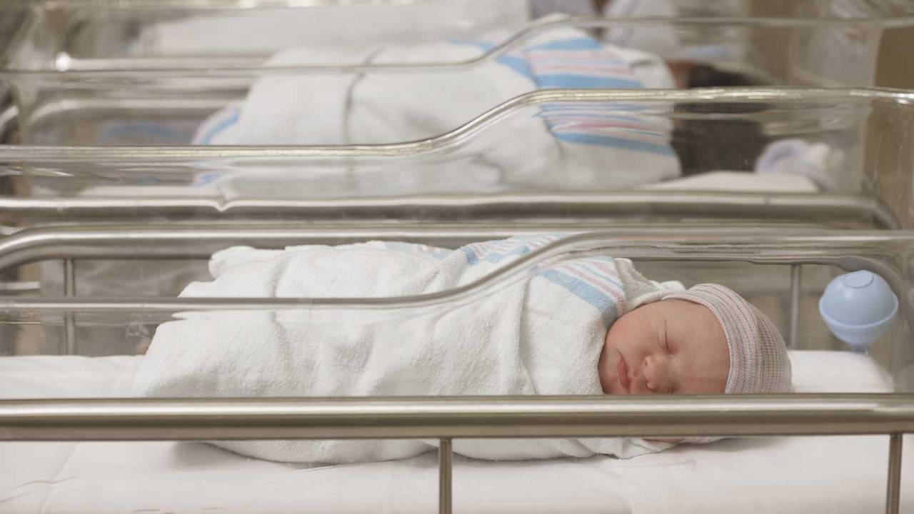 berçário de uma maternidade com vários berços e bebês dentro. Vemos uma dessas crianças de frente, enrolada em uma coberta e de touca