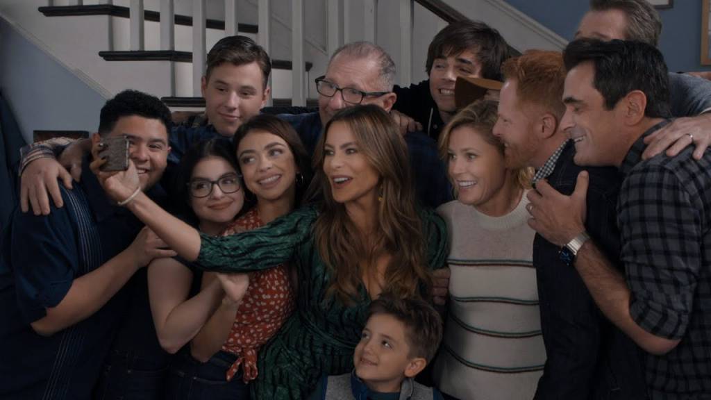 Modern-Family Cena da série. Família na cozinha, pai e filho adolescente de pé, segurando um quadro com a foto da mãe. Ela está sentada vendo, e as filhas, duas meninas adolescentes, estão sentadas de frente para ela.