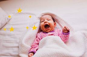 Bebê de macacão rosa, pele branca, deitado e coberto com manta branca. ELe está sorrindo com a boca bem aberta. Ao lado, há uma ilustração de constelação de Libra amarela.