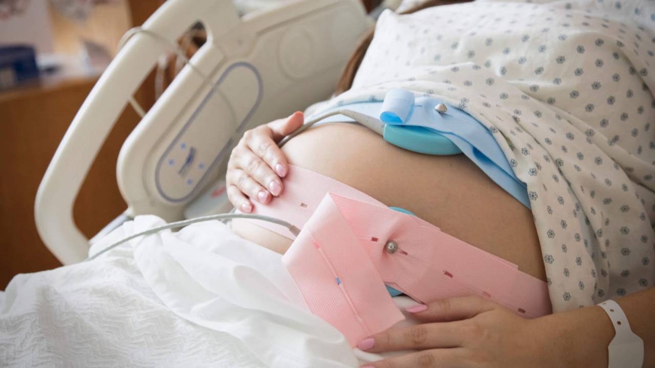 Mulher grávida no hospital com duas faixas de cardiotoco na barriga, uma azul e outra rosa.