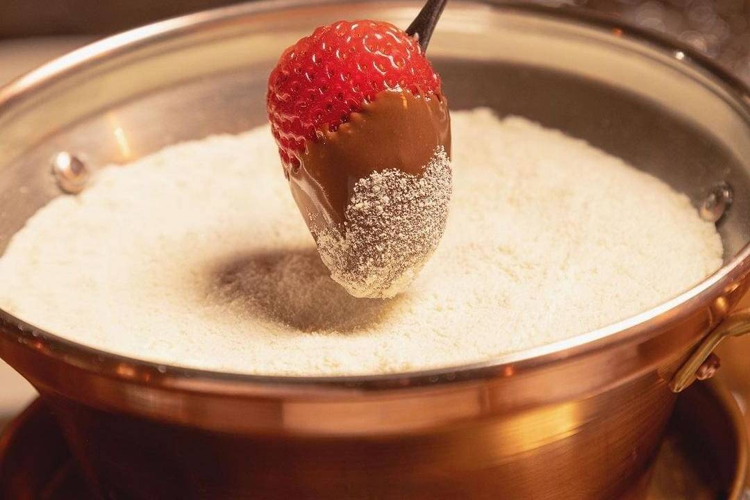 Na foto, um morango está espetado coberto por Nutella e leite em pó, pronto para ser mergulhado em uma panela de fondue, também de leite ninho. O conteúdo é claro e a panela é cobre.