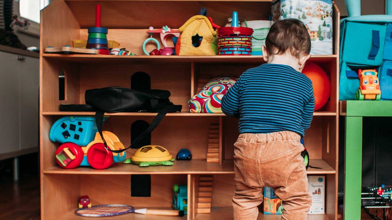 menino, de costas, mexendo em um móvel formato por prateleiras onde há brinquedos