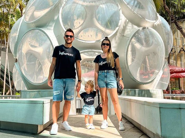 fabi-santina-entrevista-viagem-em-familia casal e criança em pé, de mãos dadas, diante de uma estrutura formada por várias outras estruturas redondas. Eles usam roupas iguais, short jeans e camiseta, olham para a frente e sorriem