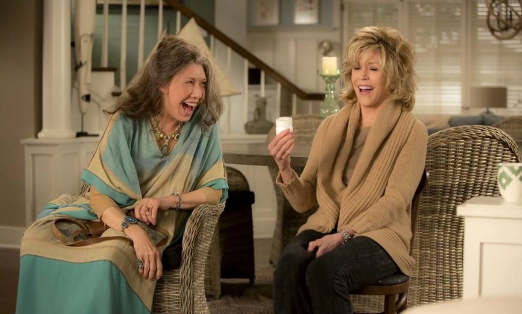 grace and frankie Cena de Grace and Frankie em que elas estão sentadas uma ao lado da outra e rindo. São duas senhoras.