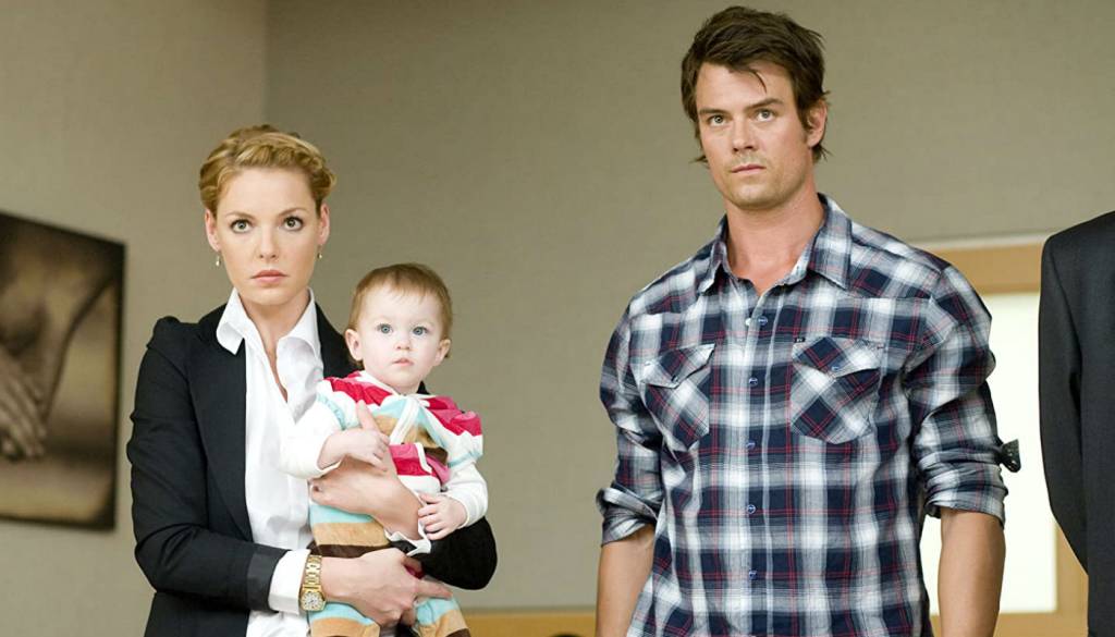 Juntos pelo acaso Cena do filme: a personagem feminina está com a bebê no colo, o personagem masculino está ao lado dela. Estão todos olhando sérios para frente.