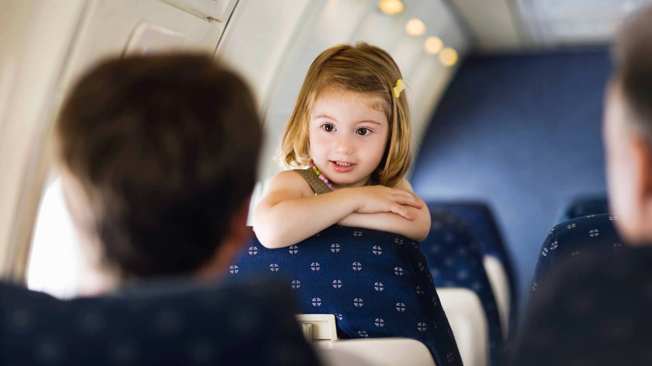 Foto dentro de avião, com uma menina loira, com por volta de três anos, virada para as poltronas de trás.