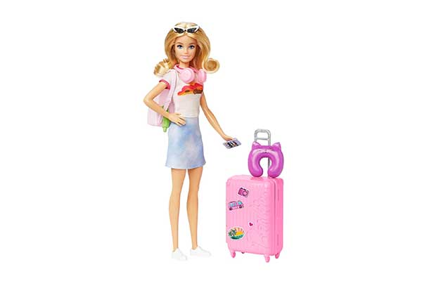 boneca Barbie com óculos de sol no cabelo e mochila nas costas, ao lado de uma mala de viagem de rodinhas, com uma almofada de pescoço em cima