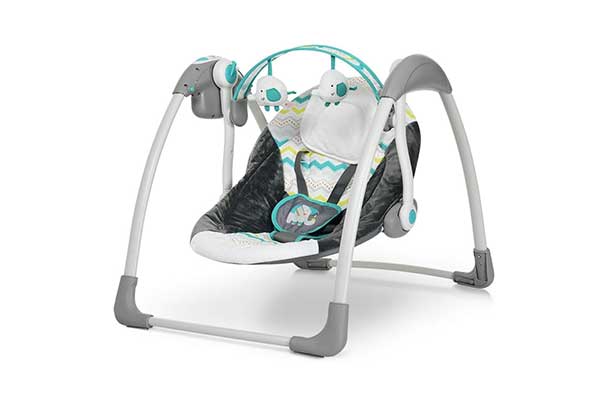 Cadeira de balanço para bebê. É cinza e branca, com estampas coloridas e geométricas no assento. Possui um arco, que fica posicionado acima do bebê, onde estão pendurados elefantes de pelúcia.