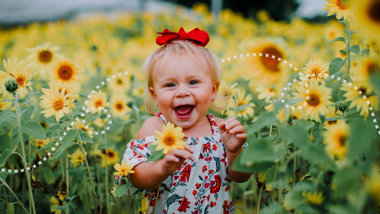 Menina sorridente segurando uma flor. Ao fundo um campo de girassóis